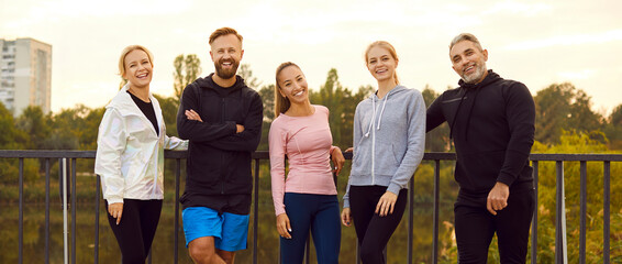 Portrait of a happy sporty people friends in sportswear looking at camera standing in a row on the bridge after successful workout in park. Outdoors training and fitness in nature concept. Banner. - obrazy, fototapety, plakaty