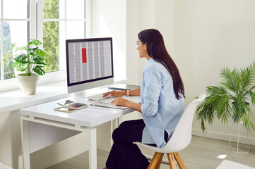 Portrait of a busy young confident business woman accountant sitting back at the desk in glasses working on a pc computer with tables and charts at modern office and typing. Accounting job concept.