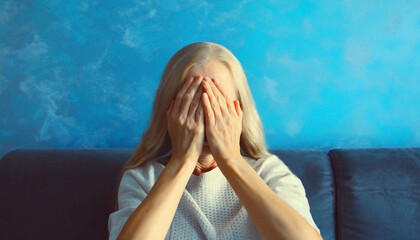 Upset sad crying middle-aged woman covering her eyes and face with her hands while experiencing mental or physical suffering sitting on couch in living room at home - obrazy, fototapety, plakaty