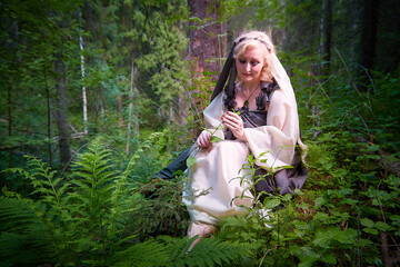 Fototapeta na wymiar Adult mature woman 40-60 in a green long fairy dress in forest. Photo shoot in style of dryad and queen of nature. Fairy who loves nature in beautiful green summer forest. Concept of caring for nature