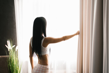 Brunette woman stands at the bedroom window in the morning. She has parted the curtains on the window and is looking at the street