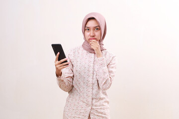 Naklejka premium Confused young asian muslim girl standing while holding a cell phone. Isolated on white