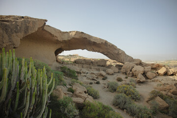 A stunning natural rock arch in a desert landscape, surrounded by arid vegetation and rocky terrain under a clear sky.