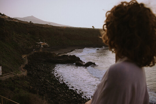 A woman with curly hair gazing at a coastal landscape during sunset, evoking feelings of solitude and reflection.
