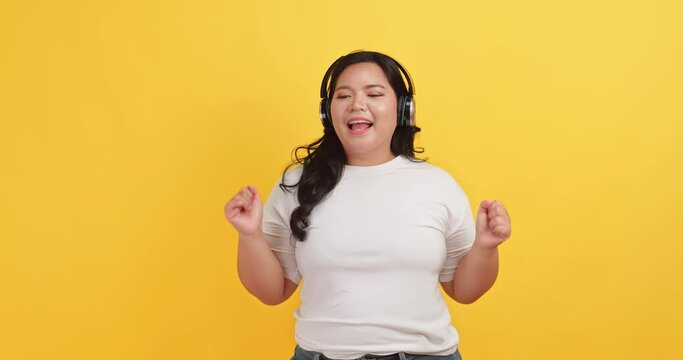 A chubby Asian woman wearing a headphones is having fun dancing on a yellow background.