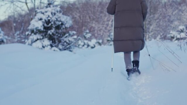 a young woman in winter in a long jacket and sunglasses walks along a snowy road among fir trees in the snow with Nordic walking poles. feet in uggs close-up
