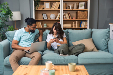 Cheerful millennial mixed couple spending time with laptop and book at home, happy young interracial lovers sitting on sofa with computer, freelance working or shopping online together and reading