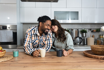 Multi-ethnic couple standing with tea cups in the kitchen and looks at each other with a love. African black man and a caucasian woman enjoy morning coffee together at home talking about life - Powered by Adobe
