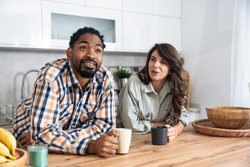 Multi-ethnic couple standing with tea cups in the kitchen and looks at each other with a love. African black man and a caucasian woman enjoy morning coffee together at home talking about life