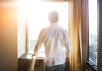 Rear view of young man looking at dawn city scenery in window after waking up.