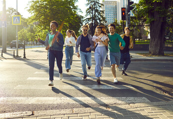 Happy multinational group of friends run across pedestrian road at red light. Attractive young people have fun on walk around the city on sunny summer day. Concept of youth lifestyle, friendship.