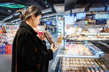 A young Asian girl is shopping in the food section of a supermarket, reading the nutritional labels of fresh baked desserts