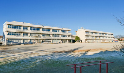 The exterior of a Japanese elementary school building and the empty dirt playing field in front of it on a cold January afternoon. 