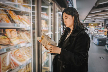 A young Asian girl is shopping for frozen noodles on a supermarket refrigerated shelf and placing...