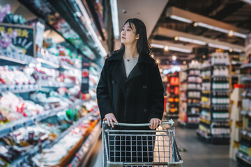 A young Asian woman is pushing a shopping cart in the food section of a supermarket, reading the nutritional labels of fresh food