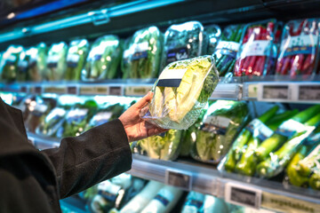 A young Asian woman with a shopping basket, standing next to a supermarket shelf, filming the purchase of fresh organic vegetables