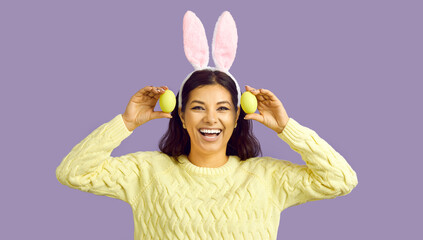 Happy Easter. Cheerful funny woman with bunny ears and with easter eggs on pastel purple background. Happy beautiful woman holding two yellow Easter eggs and sincerely smiling at camera. Banner.