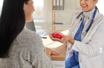 Portrait of smiling friendly doctor cardiologist holding red heart in her hands sitting at the desk...