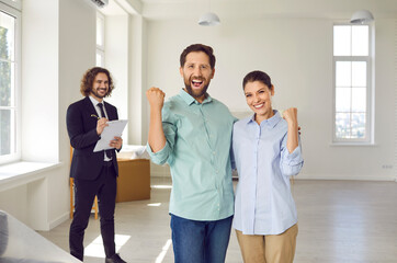 Portrait of a young excited smiling family couple happy about buying a new apartment standing with man realtor in new house making yes gesture enjoying real estate purchase. Moving day concept.