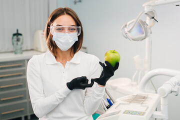 A female dentist holds a fresh apple in her hand against the background of a dental office. Women doctors. Healthy and white teeth.