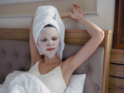 Young woman lying on bed with moisturizing beauty face mask and white towel on her head after shower, lifestyle home skincare, smile.
