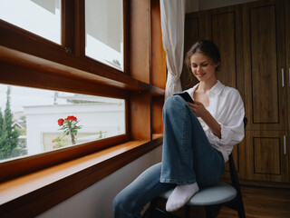 Beautiful woman with phone in hand sitting at the window with wooden frame of the house, home comfortable lifestyle with online work, cozy atmosphere and aesthetics of warmth, spring time