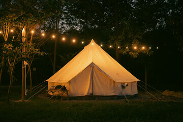 Night and warm light exposure of an illuminated bell tent surrounded by trees and a small camp fire...