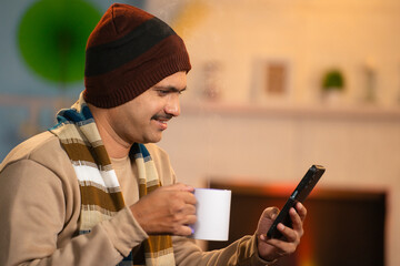 Middle aged Indian man with warmth winter wear using mobile phone by drinking tea at home - concept...