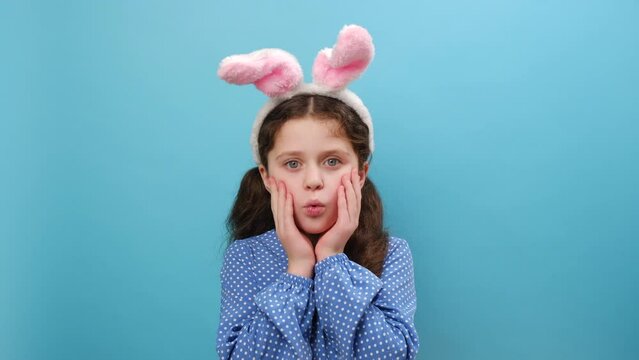 Portrait of preteen girl kid wearing cute easter bunny ears, afraid and shocked looking at camera with surprised expression, fear and excited face, posing isolated over blue color background in studio