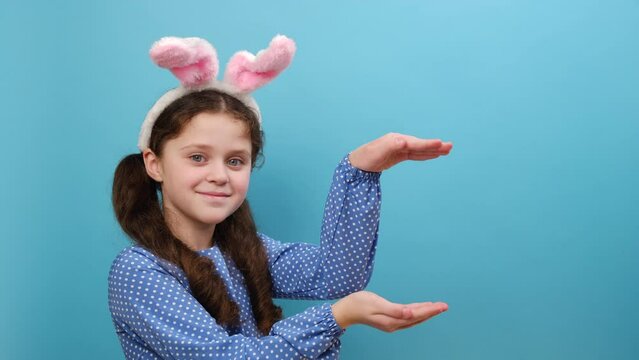 Portrait of beautiful preteen girl kid wearing cute easter bunny ears amazed and smiling to camera, raises palms as if showing something, gestures over blank space, isolated on blue studio background