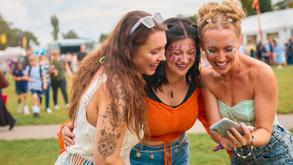 Three Female Friends Wearing Glitter Looking At Mobile Phone At Summer Music Festival