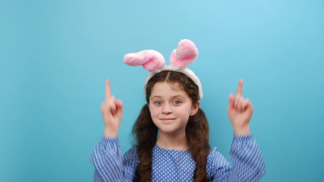 Portrait of smiling beautiful preteen girl kid wearing pink fluffy easter bunny ears, surprised looking up and pointing with fingers and raised arms, posing isolated over blue color background wall