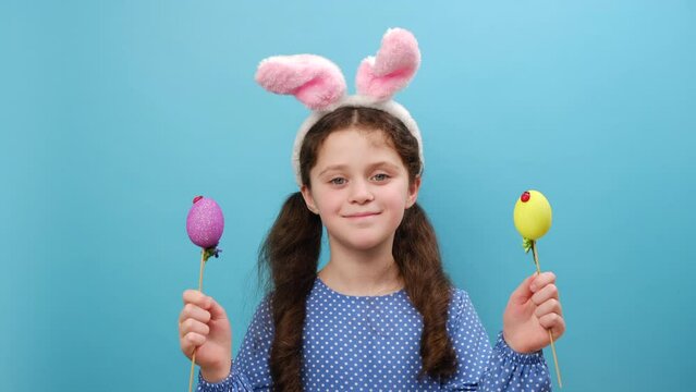Portrait of surprised smiling adorable preteen girl kid wearing fluffy bunny ears and holding colorful easter eggs, posing isolated over blue color background wall in studio. Easter holiday concept
