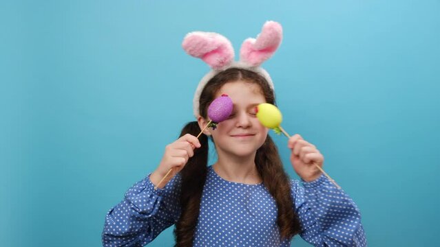 Portrait of playful funny happy preteen girl kid wearing fluffy easter bunny ears, holding near face colorful Easter eggs, posing isolated over blue color background in studio. Happy Easter concept