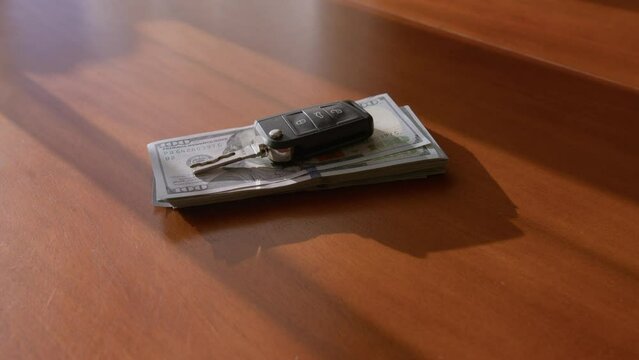 A man's hand puts one hundred dollar bills on a wooden brown table. A woman's hand rests a car key on top of them. The keychain is taken away and the money too. The concept of buying a vehicle