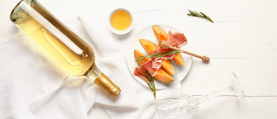 Plate with delicious melon, prosciutto, rosemary, honey and bottle of wine on white wooden...