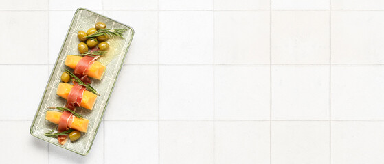 Plate with tasty melon, prosciutto, rosemary and green olives on light tile background with space for text