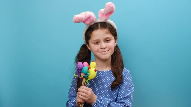 Portrait of cute positive preteen girl child in easter holiday bunny ears blowing kisses, pretty kid holding colorful eggs, happy looking at camera, posing isolated over blue studio background wall