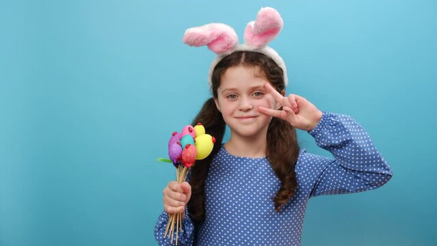 Portrait of joyful funny preteen girl kid in fluffy pink bunny ears holding toy eggs and gesturing peace sign, happy looking at camera, posing isolated over plain blue color background wall in studio