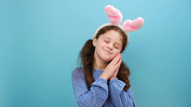 Portrait of drowsy smiling cute preteen girl kid in pink bunny ears yawning, sleepy inattentive, dressed in dress, posing isolated over blue color background wall in studio. Easter holiday concept