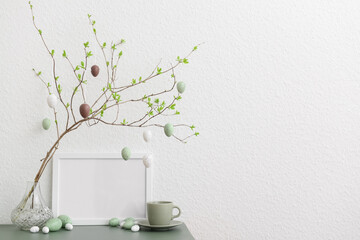 Tree branch with hanging Easter eggs, cup and blank frame on table near white wall