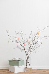 Vase with tree branches and Easter eggs in nest on boxes near white wall