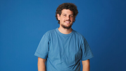 Fototapeta premium Smiling guy with curly hair dressed in blue t-shirt looking at the camera and waving his hand in welcoming gesture, saying hello to someone, isolated on blue background in the studio