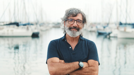 Happy male seigneur in glasses crossing his arms while standing in the port and looking at the camera on yachts background, camera is moving closer - obrazy, fototapety, plakaty