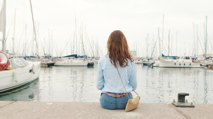 Attractive slim middle-aged woman sitting in the port looks into the distance at the yachts, back view