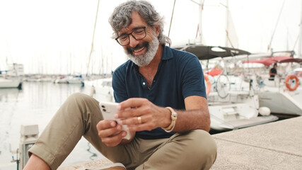 Close up, happy senior man in glasses sits in the port on yacht background uses smartphone, looks at funny video, scrolling on the phone screen, communicates in social networks