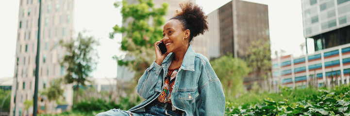 Closeup portret cute African girl with ponytail, wearing denim jacket, sits with morning coffee and makes stream, video call on modern city background. - obrazy, fototapety, plakaty