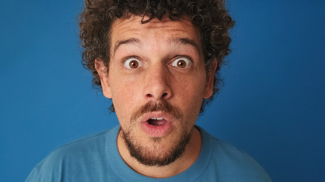 Close-up of guy with curly hair dressed in blue t-shirt, looking at the camera, looking for something, surprised with wide eyes on blue background in the studio