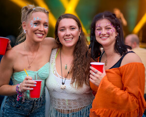 Portrait Of Three Female Friends Wearing Glitter Having Fun At Summer Music Festival Holding Drinks