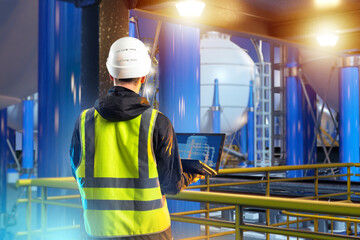 Man industrial engineer. Guy on territory of plant. Industrialist with laptop with back to camera. Oil refinery engineer. Man stands on industrial mezzanine. Industrial engineer near oil tanks - obrazy, fototapety, plakaty